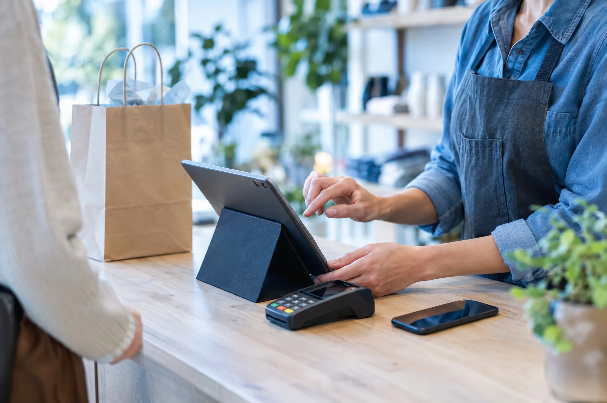 A shop owner rings up a sale on a tablet point-of-sale system beside a card reader.